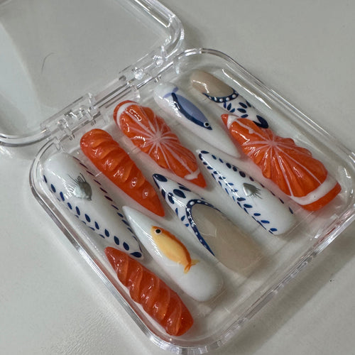 A set of decorative press-on nails in a clear plastic case, featuring orange salmon and tuna sushi designs alongside white nails with blue wave patterns and small fish details.