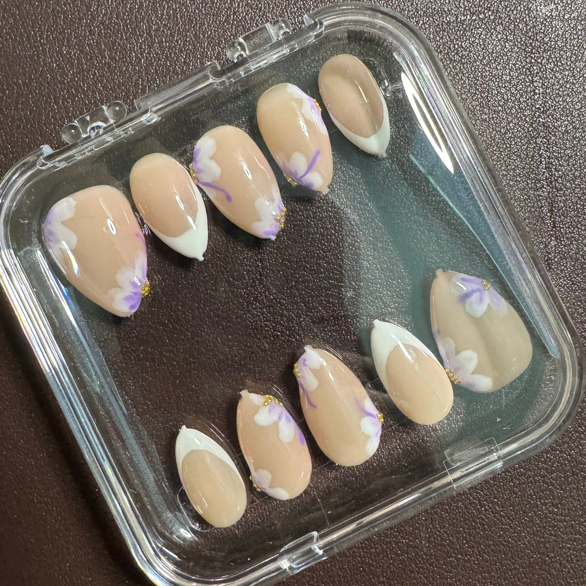 Ten press-on nails with a nude beige base, white French tips, and light purple floral designs, arranged inside a clear plastic case on a dark brown textured surface.