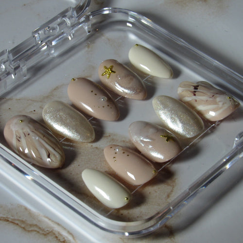 A set of press-on nails displayed in a clear plastic case on a light marble-like surface. The nails feature neutral shades including beige, cream, white, and metallic gold glitter, with some having delicate gold leaf accents.