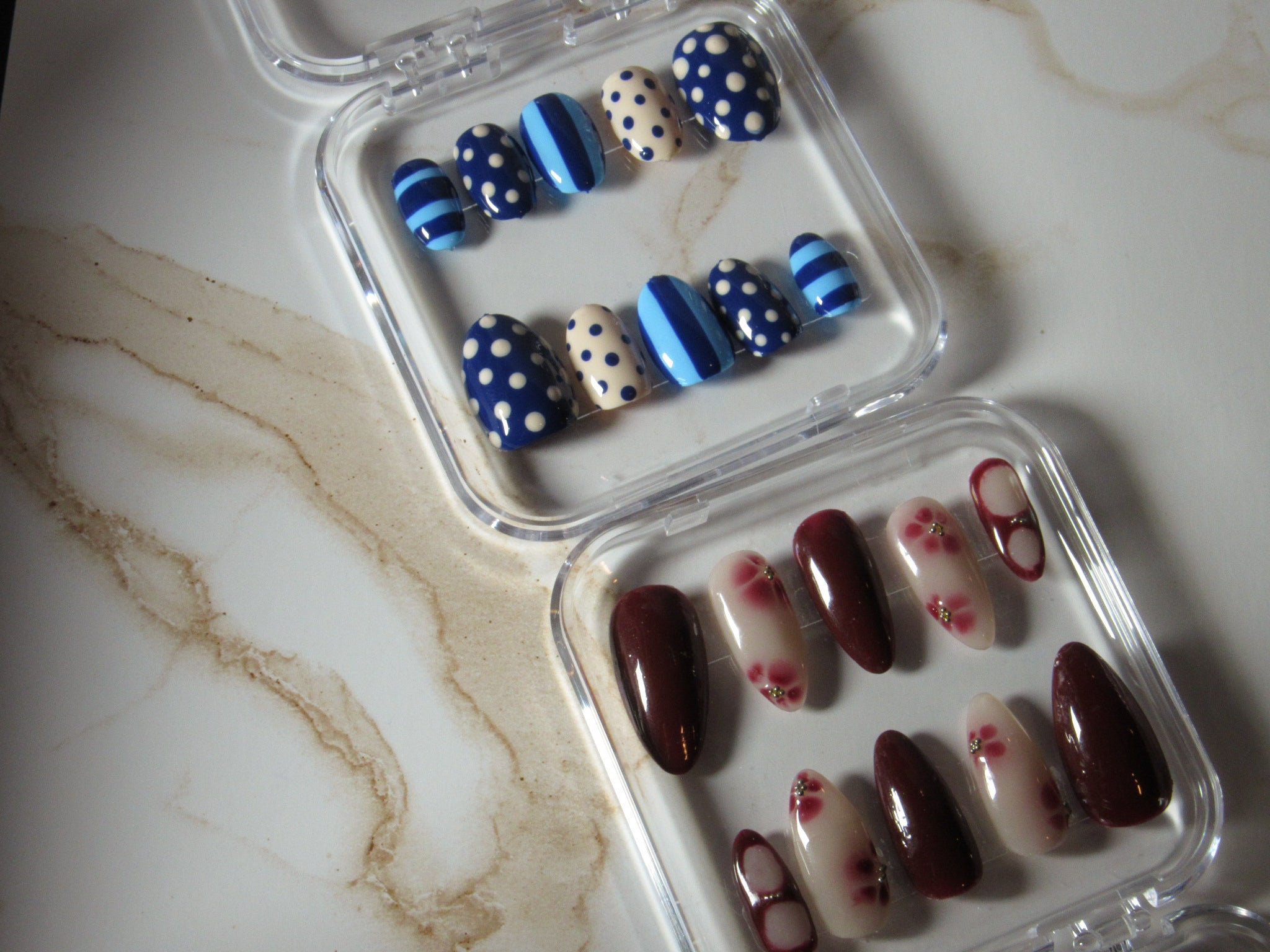 Two pairs of press-on nails in clear plastic cases, displayed on a white marble surface. One set features navy blue, light blue, and white polka dots and stripes, while the other features glossy dark burgundy and white with red floral details.