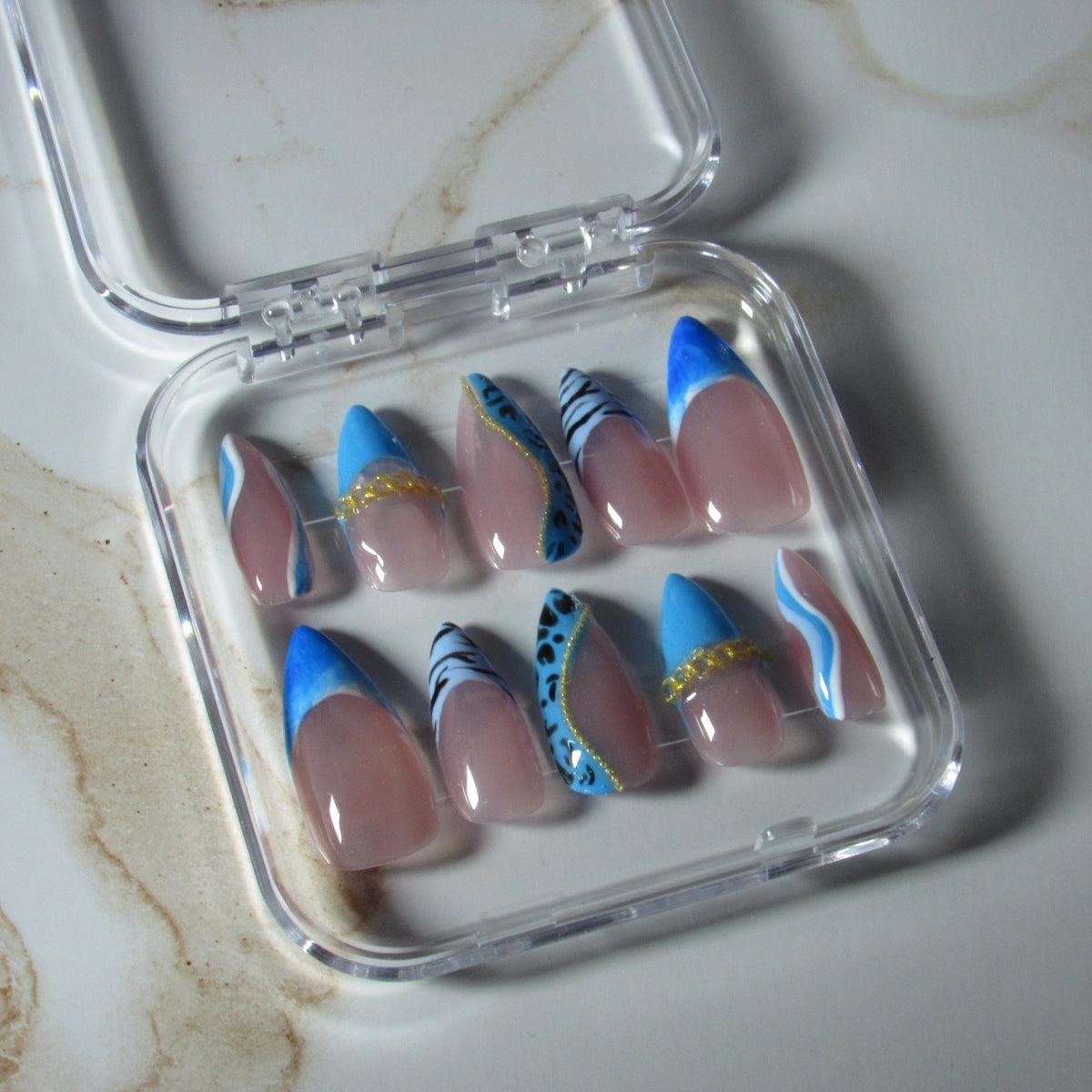 A set of false press-on nails in a clear plastic case, featuring a nude pink base with blue tips, some with gold glitter and others with blue and white patterns.