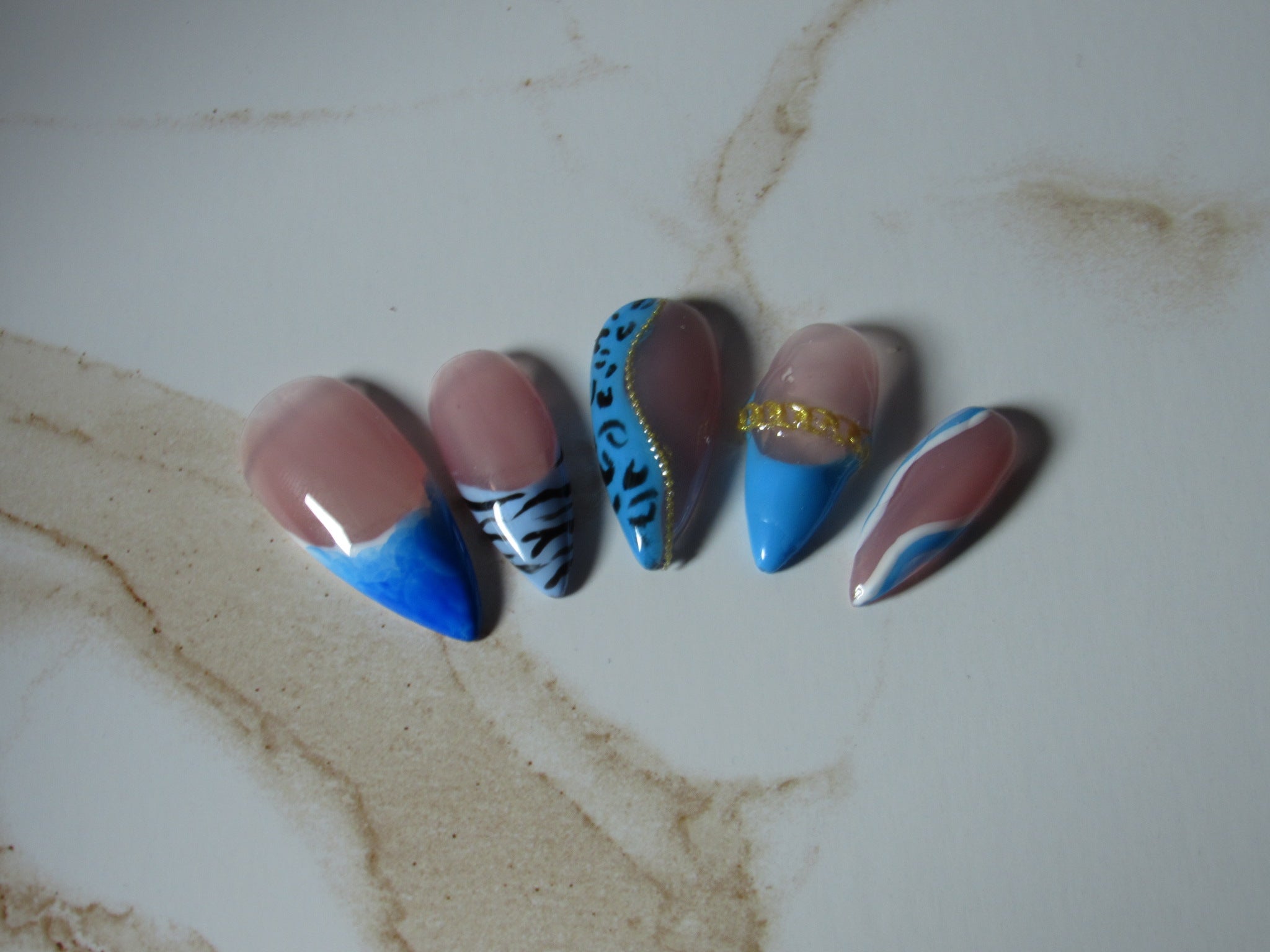 Five artificial almond-shaped nails displayed on a light marble surface, featuring designs of blue and pink French tips, black and white zebra stripes, blue leopard print with gold trim, solid blue with a gold chain, and pink and blue French tips.