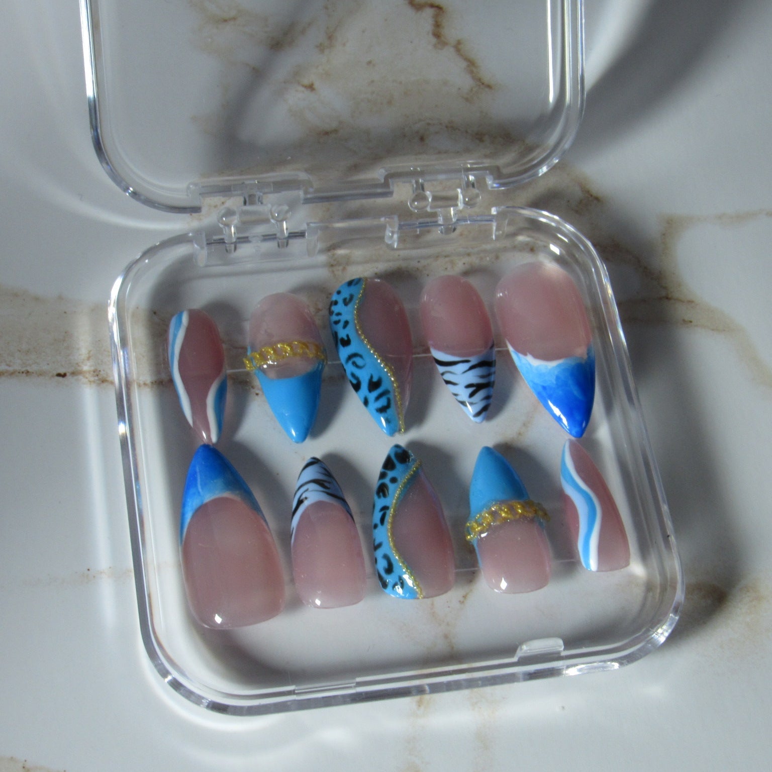 A set of 10 false press-on nails displayed in a clear plastic case on a white marble surface. The nails feature a mix of solid light pink, bright blue, black-and-white zebra stripes, leopard print, and gold glitter accents.