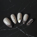 Five false press-on nails with a glossy cream-white base, featuring various decorative designs including gold dots, a small green leaf, and a black-and-white polka dot French tip, displayed on a black marble surface.