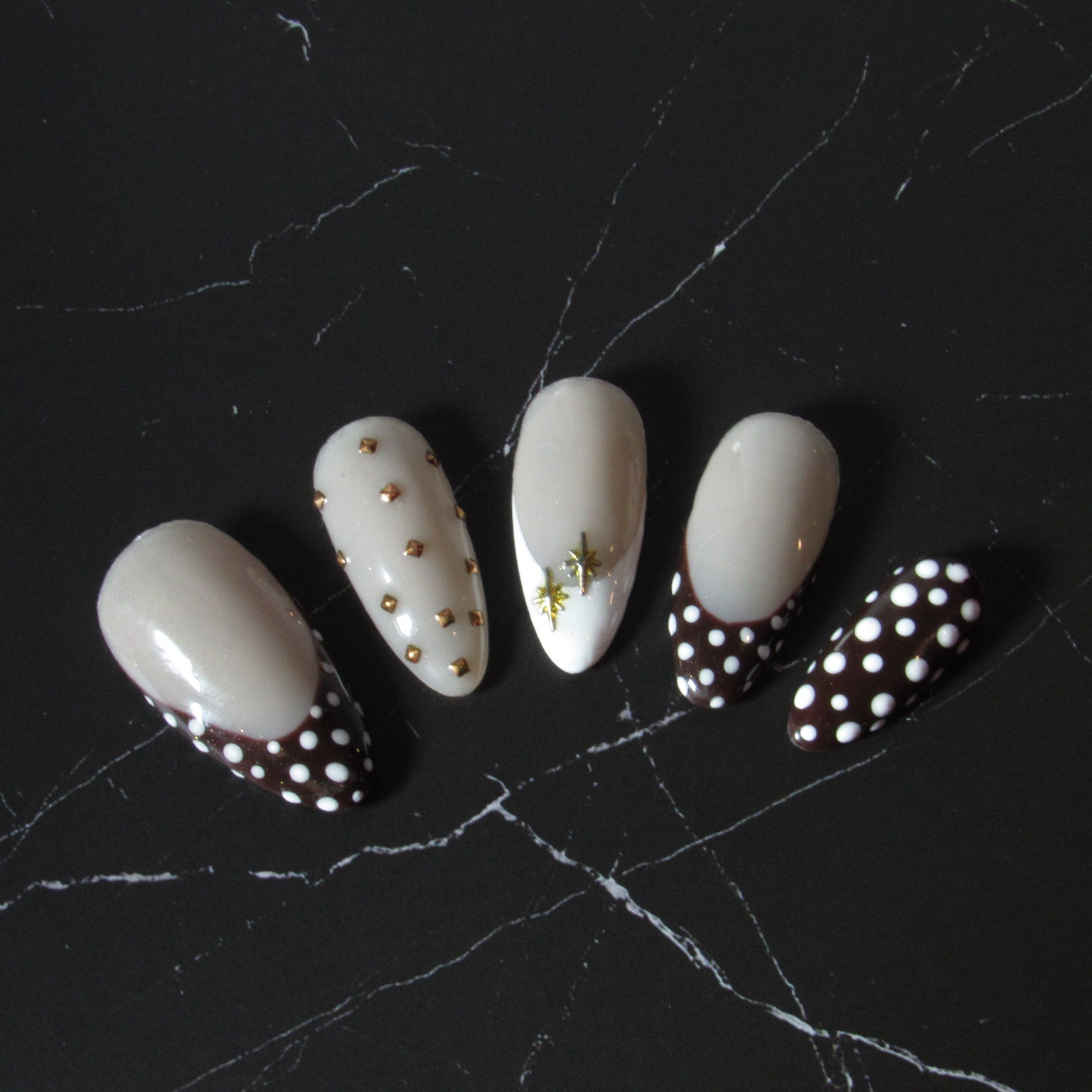Five false press-on nails with a glossy cream-white base, featuring various decorative designs including gold dots, a small green leaf, and a black-and-white polka dot French tip, displayed on a black marble surface.