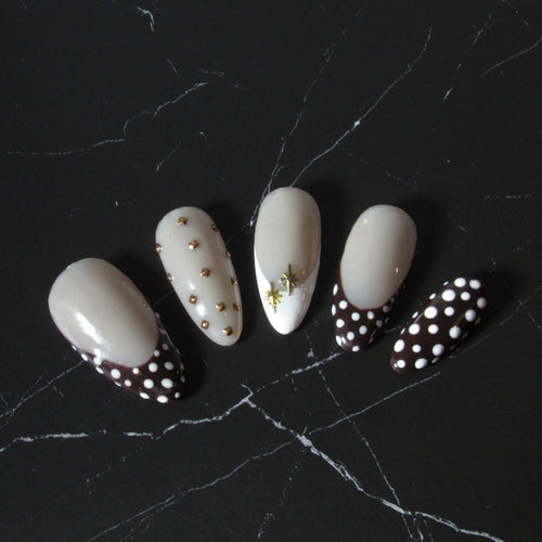 Five false press-on nails with a glossy cream-white base, featuring various decorative designs including gold dots, a small green leaf, and a black-and-white polka dot French tip, displayed on a black marble surface.