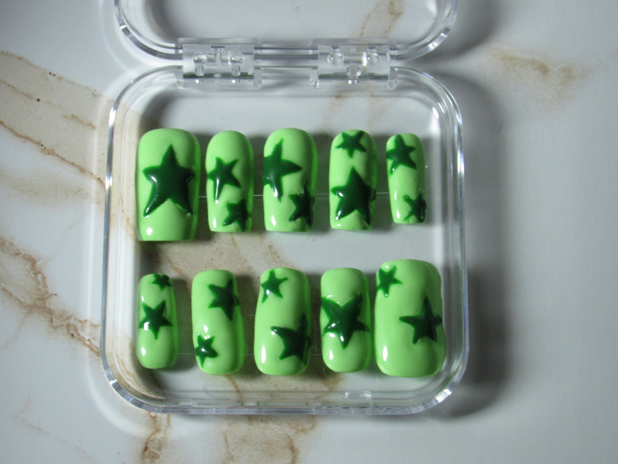 A set of ten press-on nails in a clear plastic case, featuring a light green base with dark green star patterns, displayed on a light marble surface.