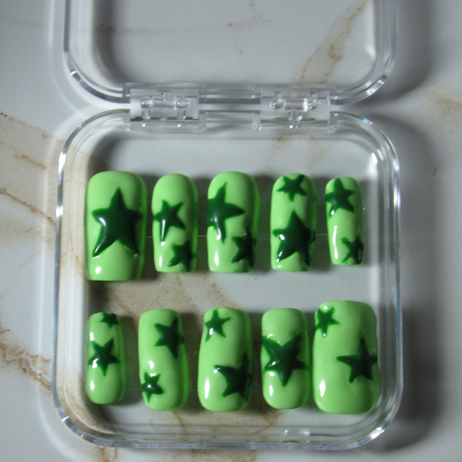 Green press-on nails with dark green star patterns, displayed in a clear plastic case on a marble surface.