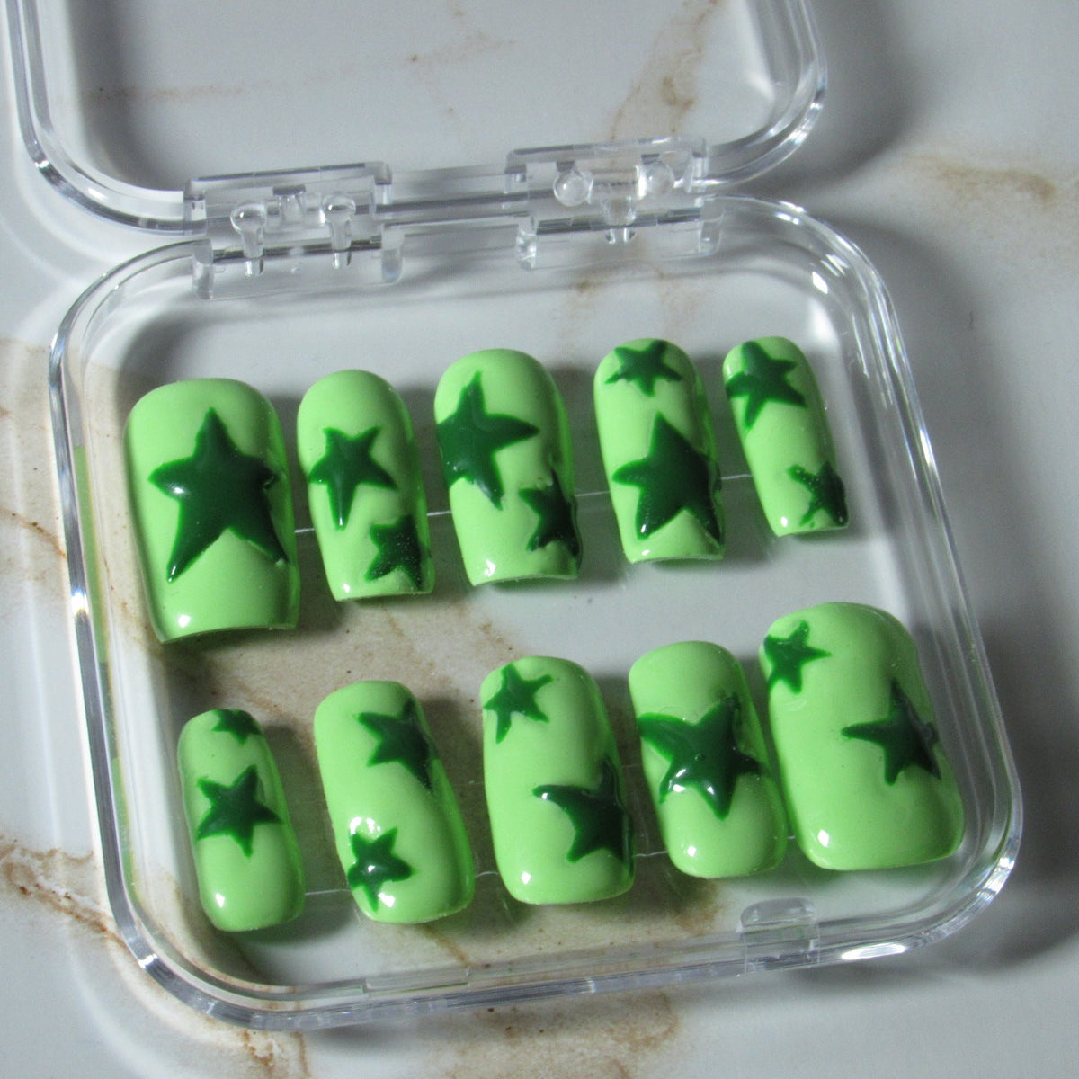 Green press-on nails with dark green star designs, displayed in a clear plastic case on a light marble surface.
