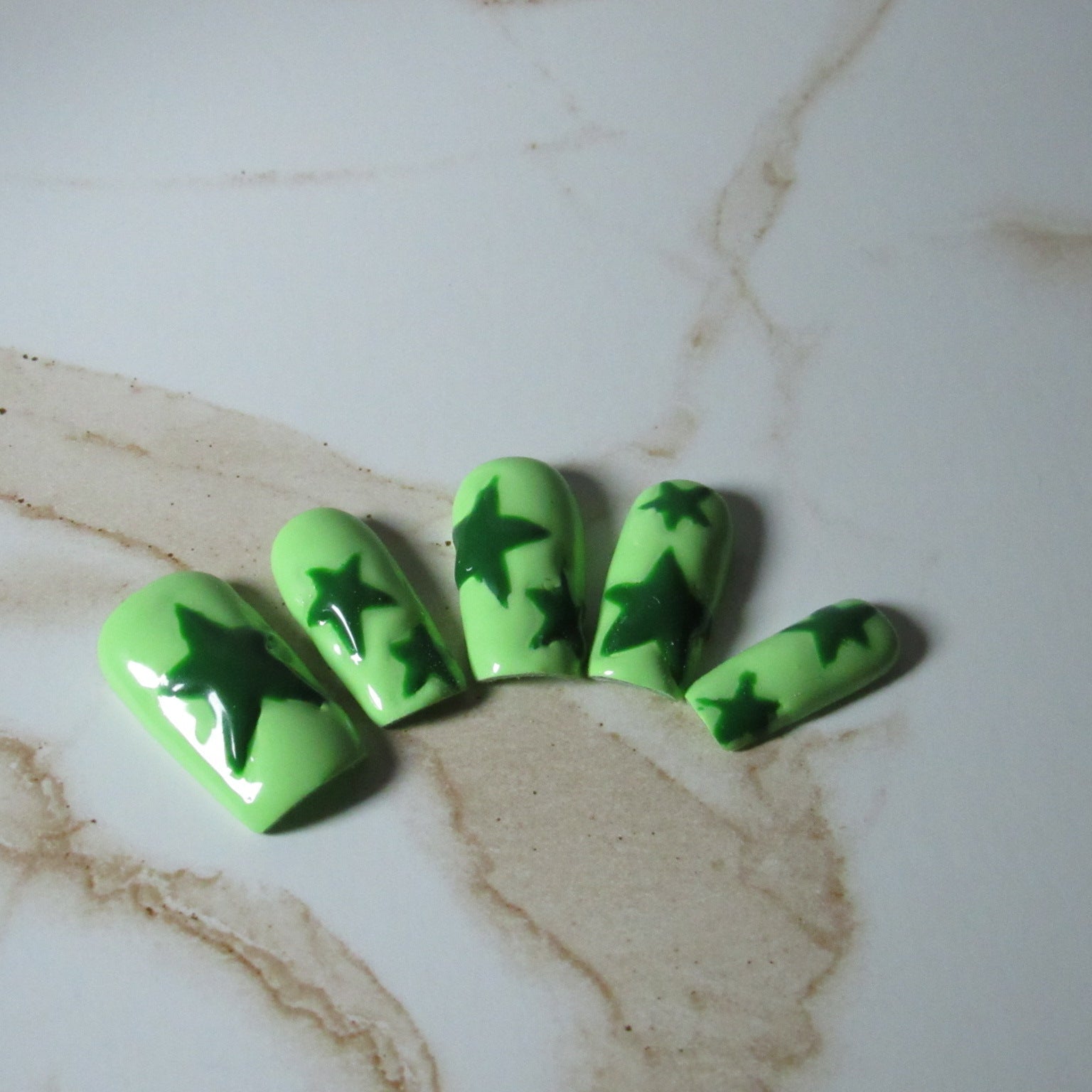 Five green press-on nails with dark green star patterns, arranged on a white and beige marble surface.