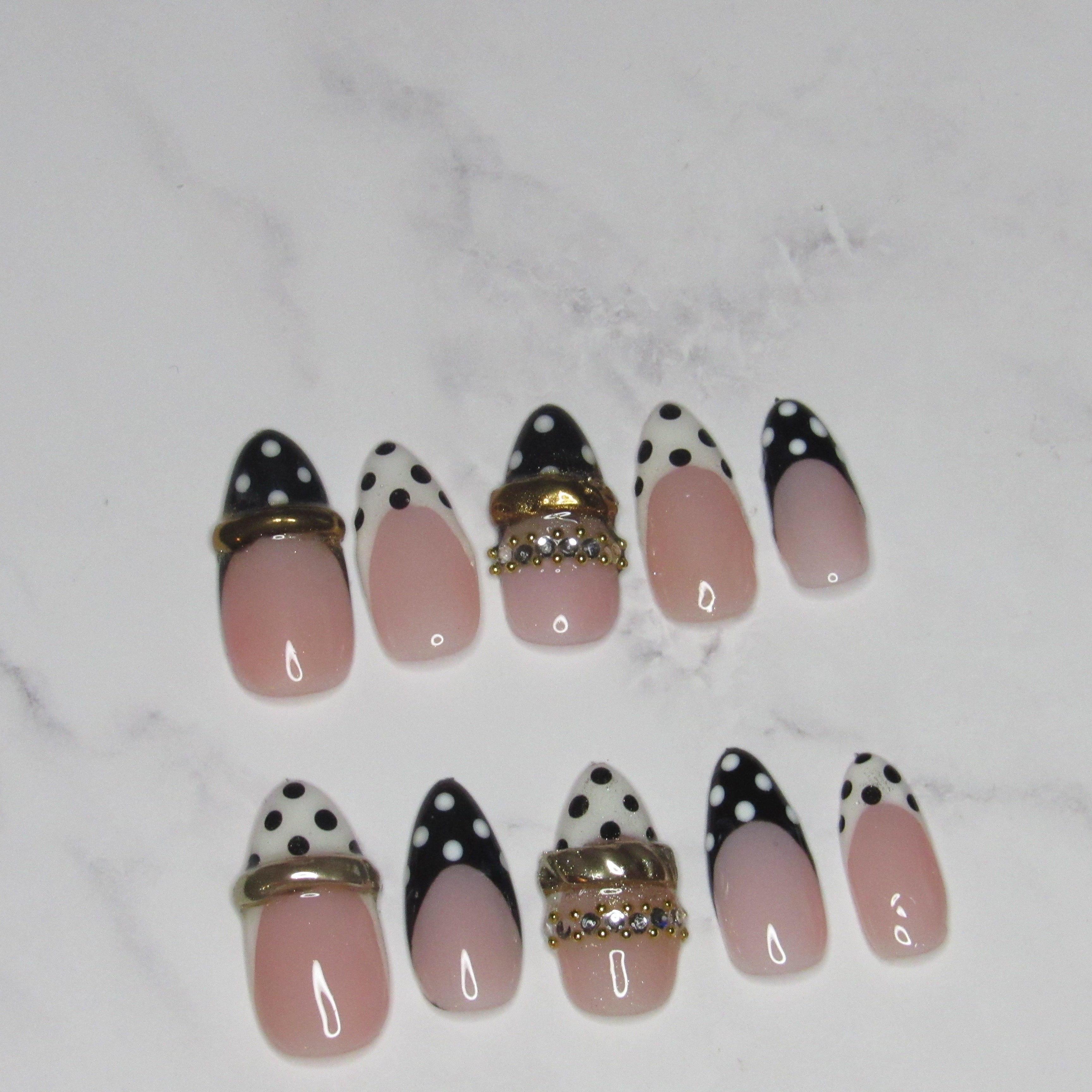 A set of artificial press-on nails arranged on a white marble surface. The nails feature a pink-to-white ombre base with black polka-dot pointed tips, some decorated with gold metallic bands and rhinestone accents.