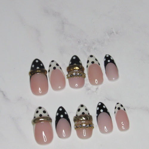 A set of artificial press-on nails arranged on a white marble surface. The nails feature a pink-to-white ombre base with black polka-dot pointed tips, some decorated with gold metallic bands and rhinestone accents.