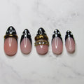 Five artificial press-on nails arranged vertically on a white marble surface. The nails feature a glossy pink base with a black French tip design, white polka dots, and one nail decorated with a gold-tone rhinestone ring accent.