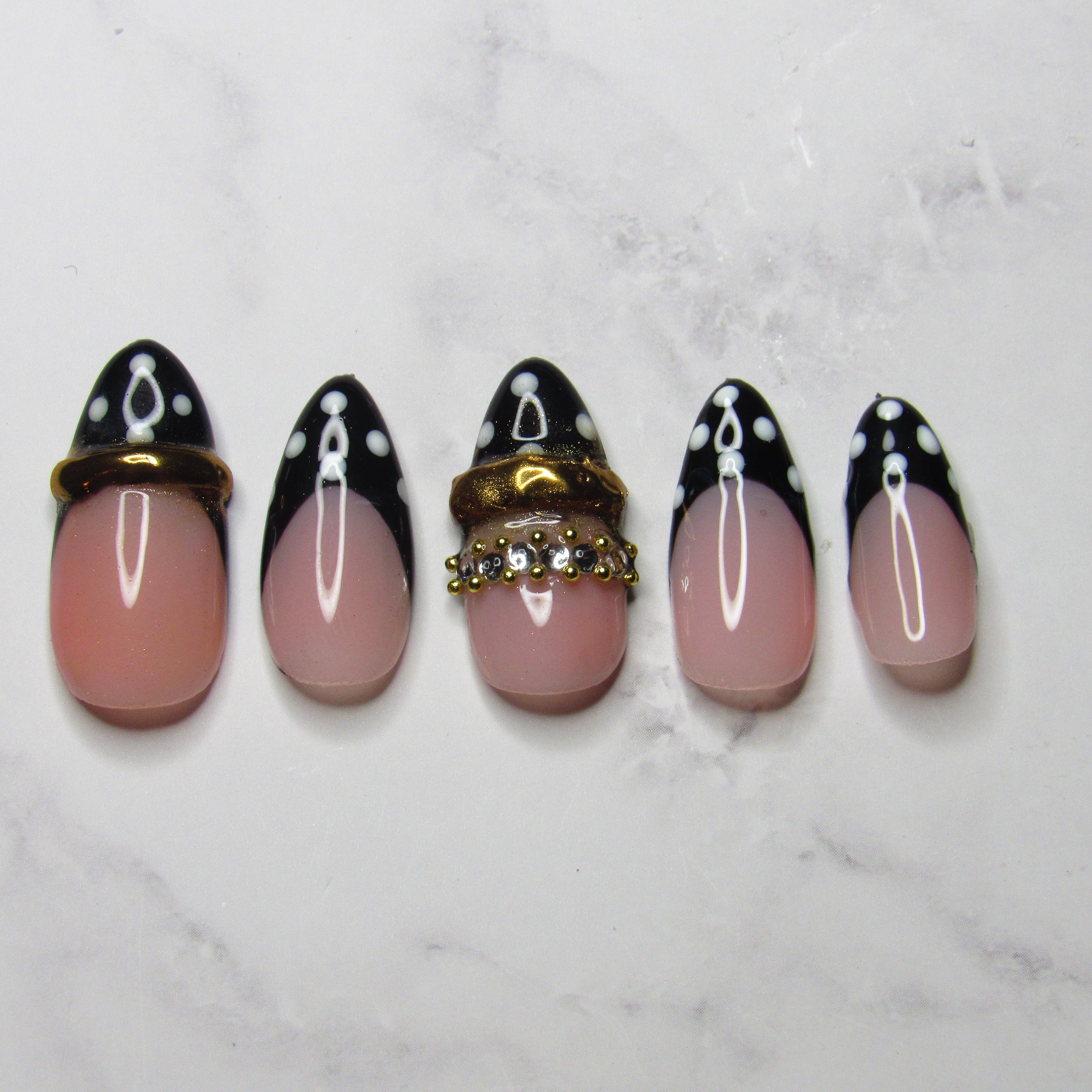 Five artificial press-on nails arranged vertically on a white marble surface. The nails feature a glossy pink base with a black French tip design, white polka dots, and one nail decorated with a gold-tone rhinestone ring accent.