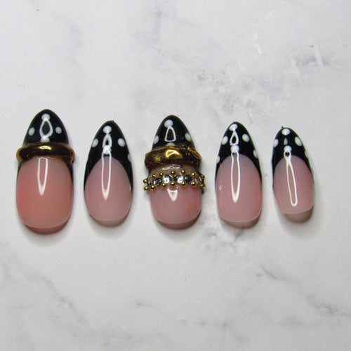 Five artificial press-on nails arranged vertically on a white marble surface. The nails feature a glossy pink base with a black French tip design, white polka dots, and one nail decorated with a gold-tone rhinestone ring accent.