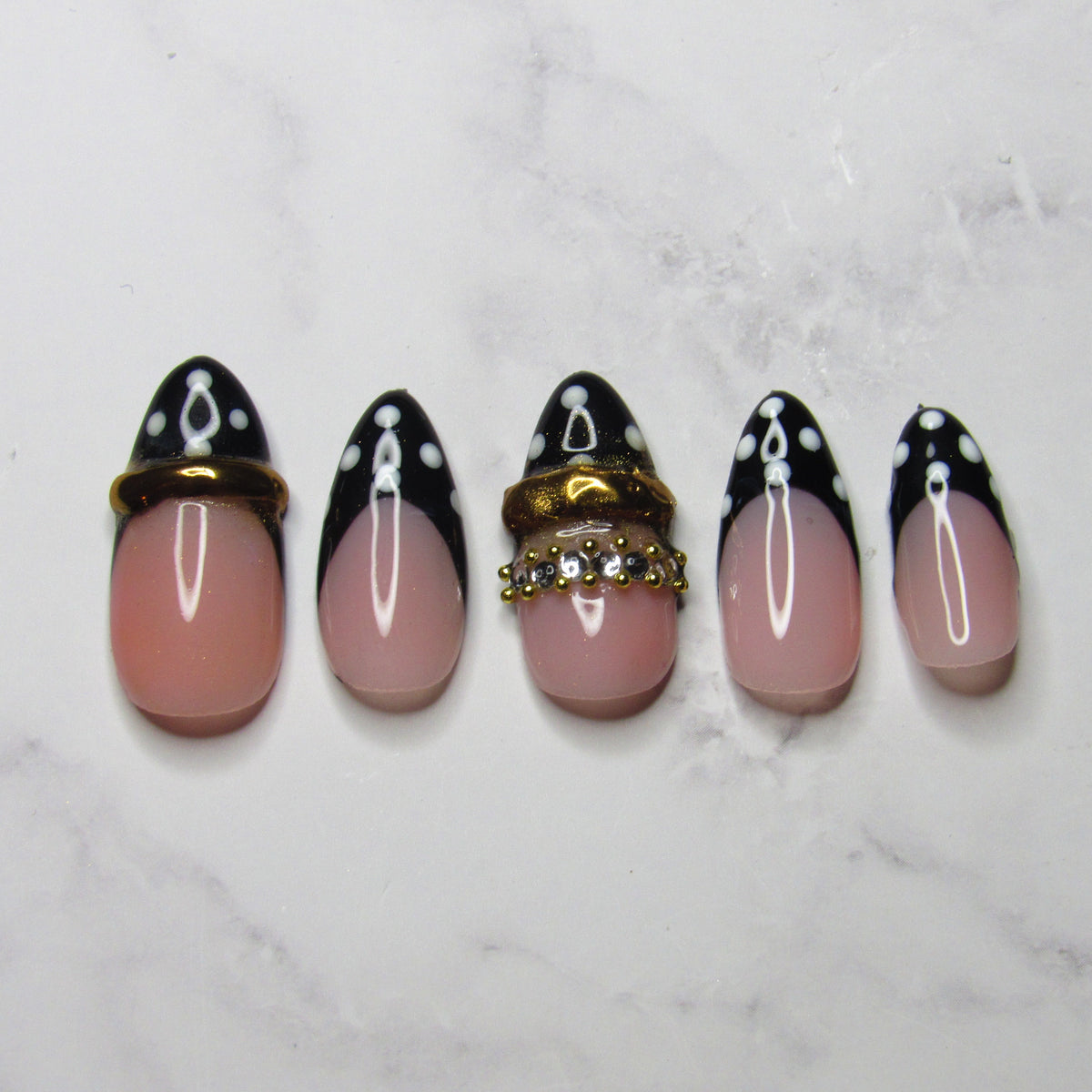 Five false press-on nails arranged vertically on a white marble surface. The nails feature a glossy pink-to-white ombre gradient on the lower half and a black-to-white ombre design with white polka dots on the upper half, with two nails decorated with gold rings and rhinestones.