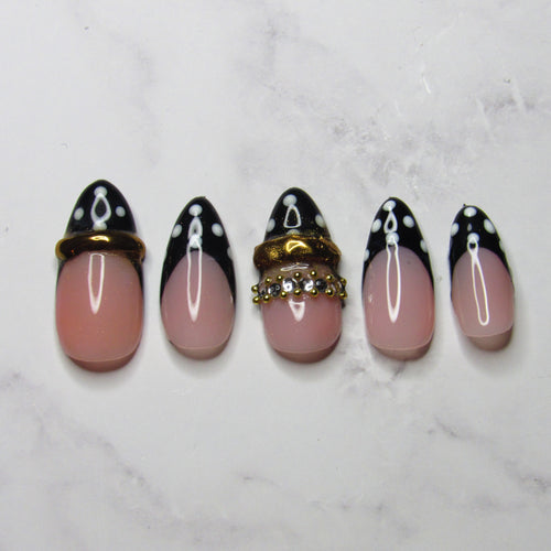 Five false press-on nails arranged vertically on a white marble surface. The nails feature a glossy pink-to-white ombre gradient on the lower half and a black-to-white ombre design with white polka dots on the upper half, with two nails decorated with gold rings and rhinestones.